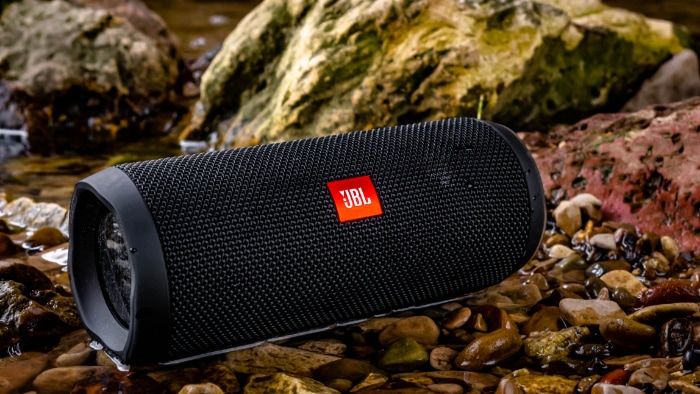 A black JBL Bluetooth speaker on rocky riverside pebbles