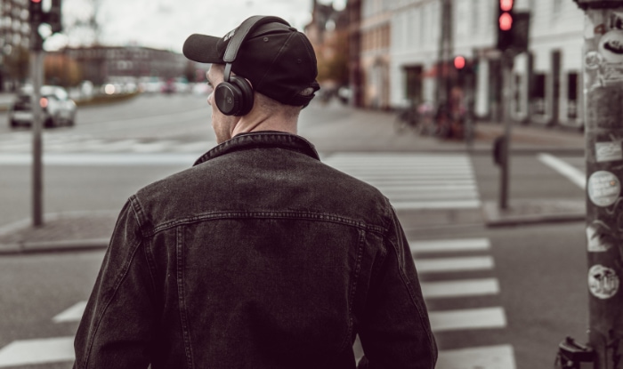 A man wearing a black cap and over ear headphones waiting to cross a city street