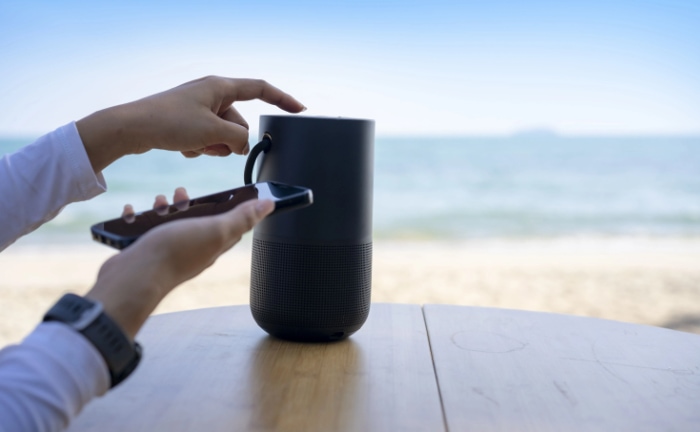 A persons hand pairing a smartphone with a Bluetooth speaker on a beachside table