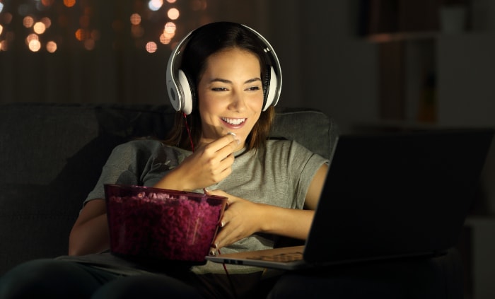 A woman holding a bowl of popcorn while using headphones and a laptop