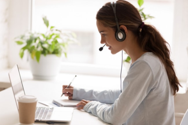 A woman using a headset and taking notes while working on her laptop