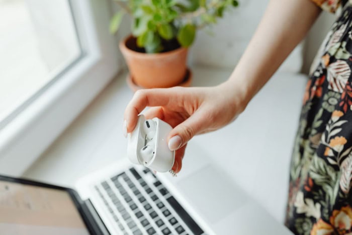 Hand holding white wireless earbuds case near laptop