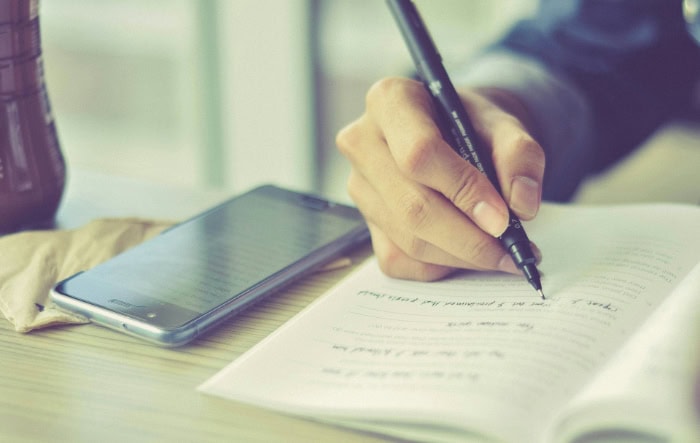 Hand writing in notebook next to smartphone on desk