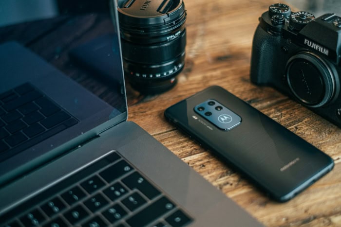 Laptop Fujifilm camera lens and Motorola smartphone on wooden table