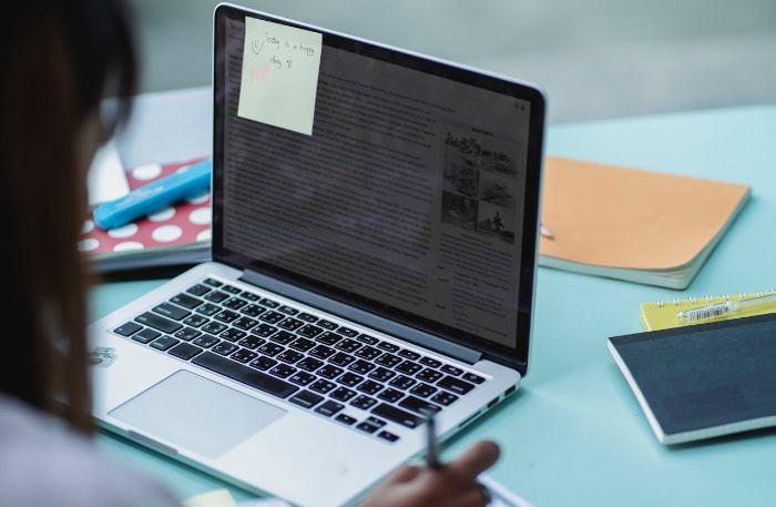 Person writing notes next to laptop on desk 1