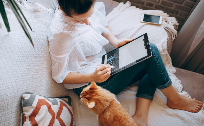 Woman with tablet and stylus on couch accompanied by orange cat