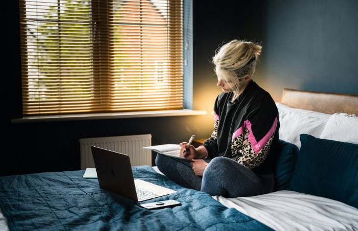 Woman writing in notebook on bed with laptop nearby